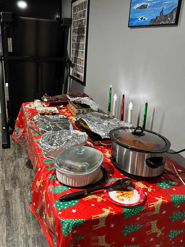 A festive holiday table adorned with a red tablecloth featuring Christmas decorations, displaying various covered dishes, a slow cooker, and lit candles.