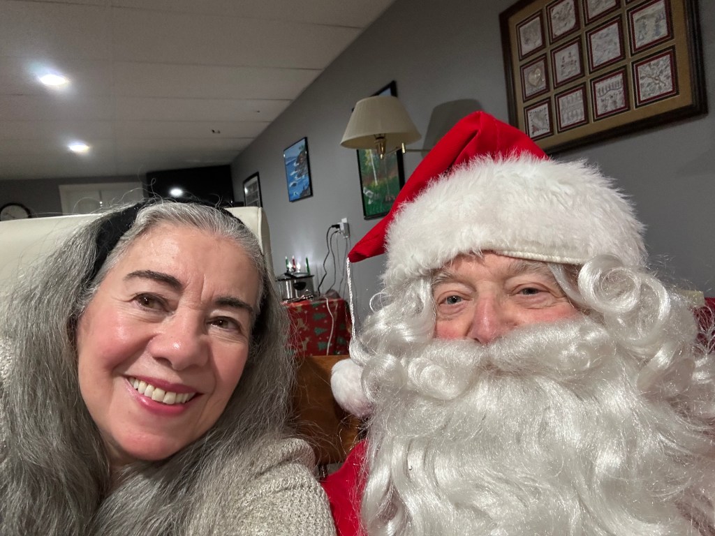 A smiling woman poses for a selfie with Santa Claus, who is wearing a traditional red suit and a white beard. They are in a festive setting with holiday decorations in the background.