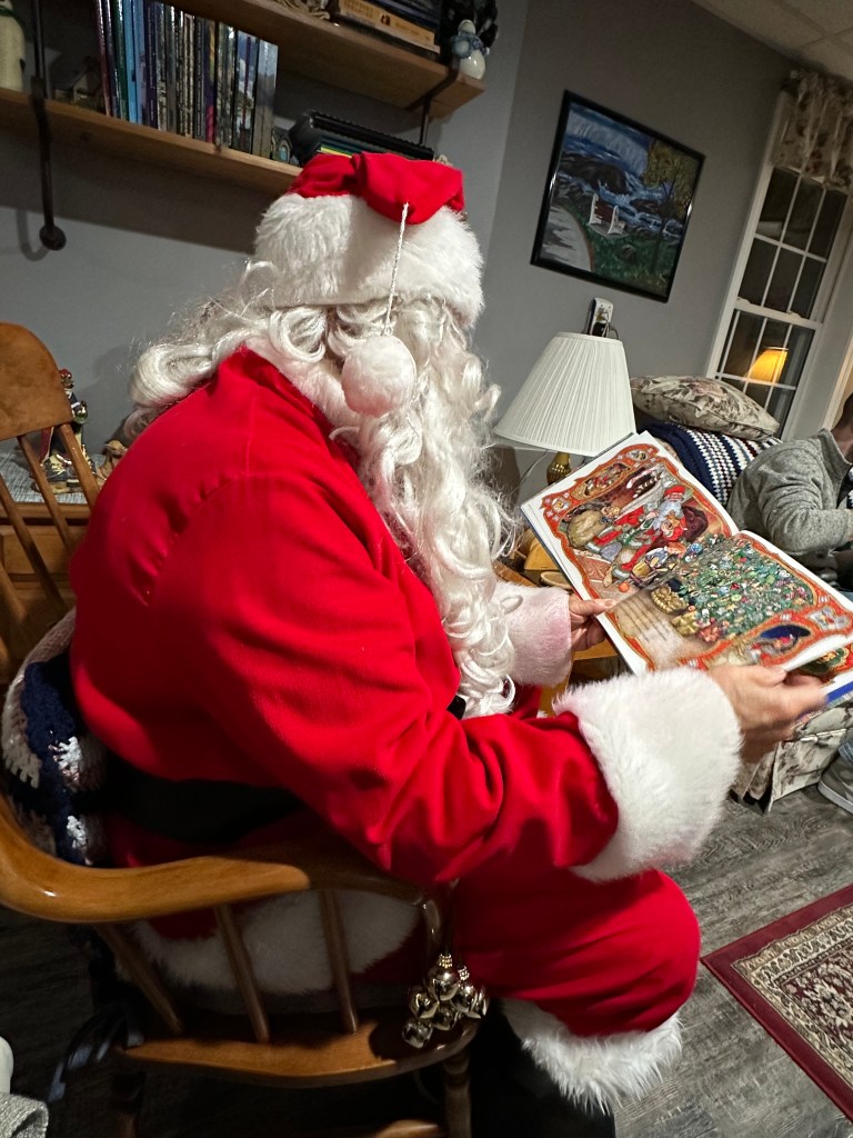 Santa Claus reading a book while sitting in a cozy living room, decorated for the holidays.