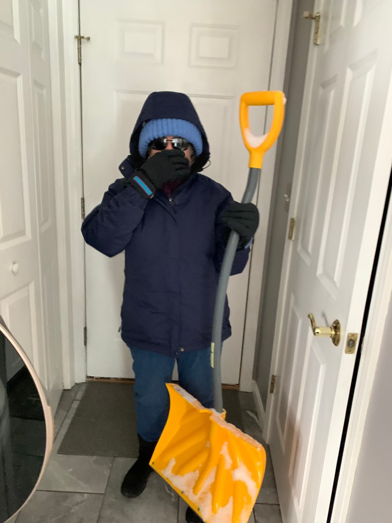 A person dressed warmly in a blue coat, hat, and sunglasses stands indoors holding a yellow snow shovel, ready to go outside.