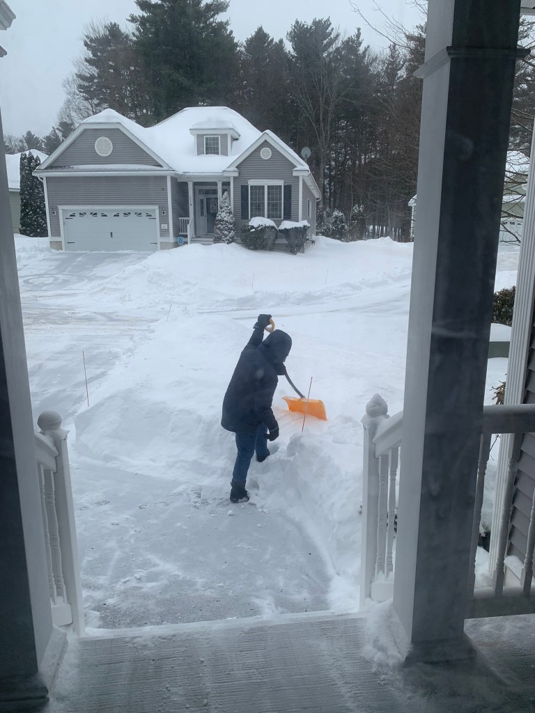 A person shoveling snow in front of a house covered in snow during winter.