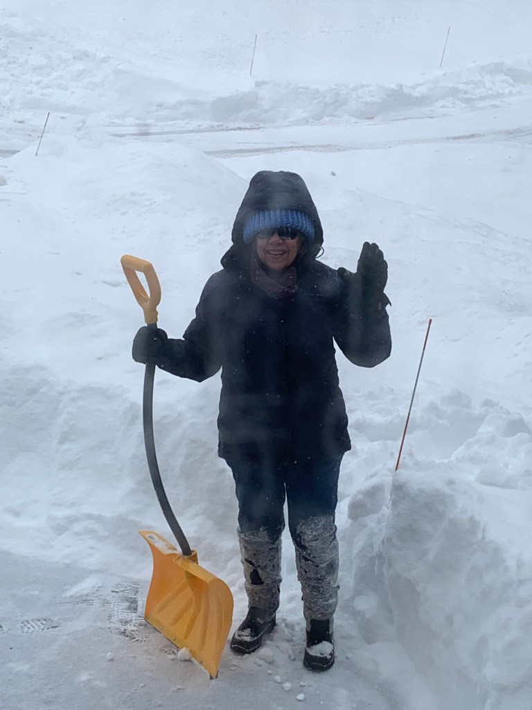 A person wearing winter clothing and sunglasses stands outside in heavy snow, holding a yellow snow shovel and waving.