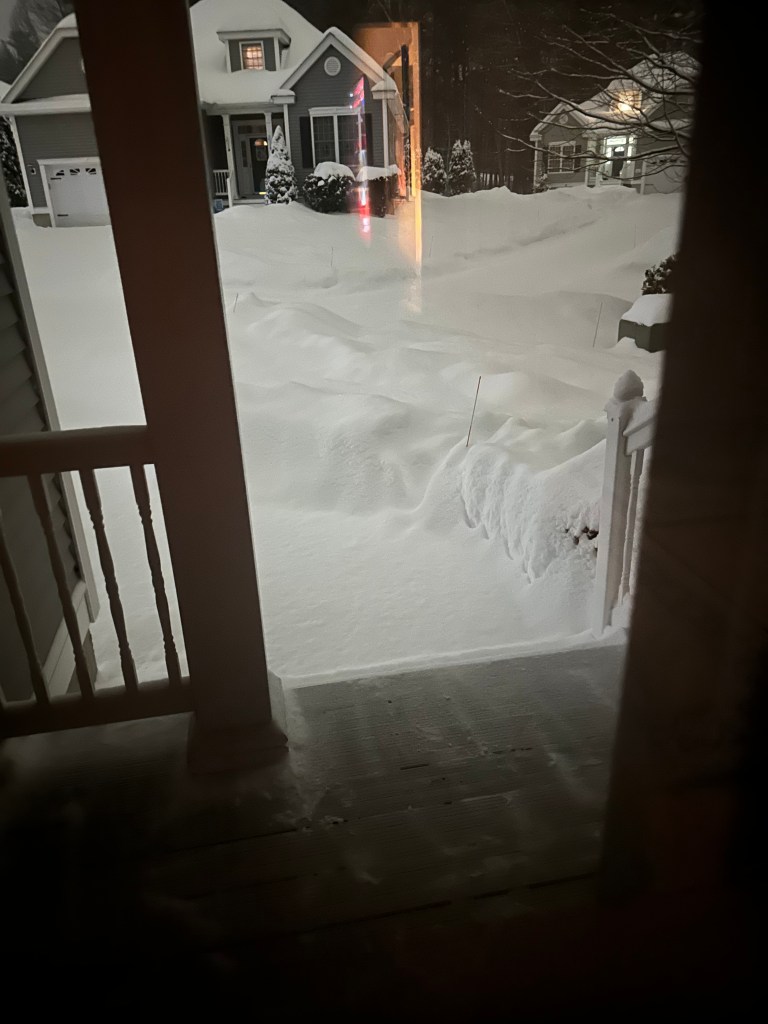 A view of a snowy front yard at night, with thick layers of snow covering the ground and surrounding the house.