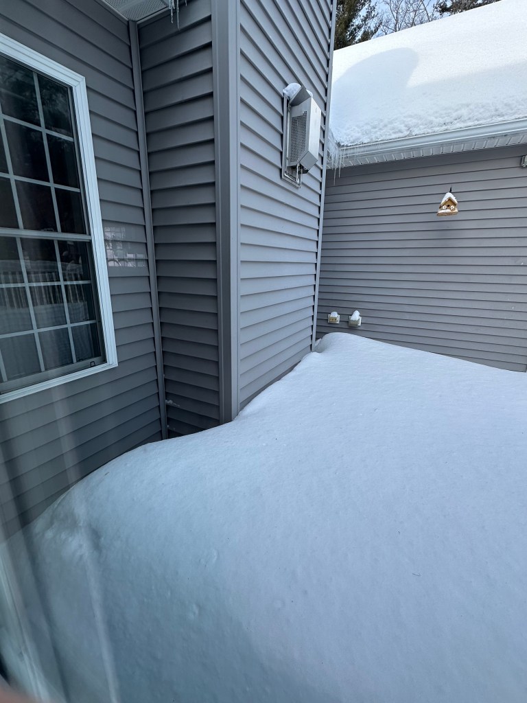 Corner of a house showing snowdrifts against the siding, with a window and an air conditioning unit visible.