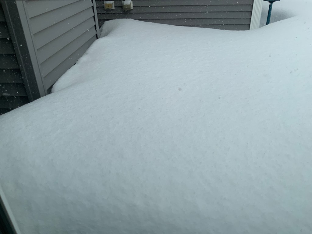 Thick layer of snow covering a surface near a house, with siding visible in the background.
