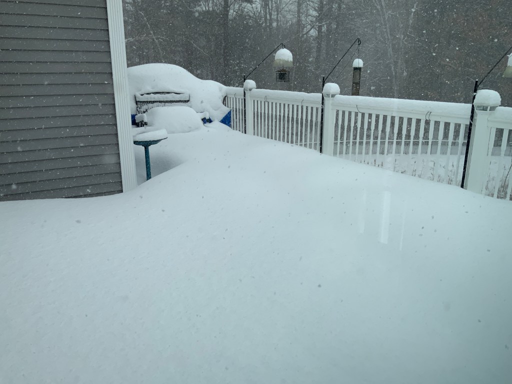 A backyard covered in deep snow during a heavy snowfall, with a grill and patio furniture partially buried, and a white fence in the background.