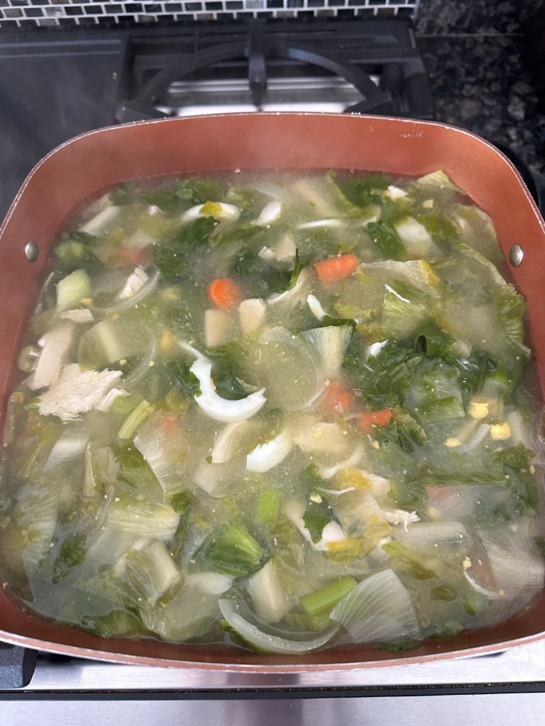 A close-up view of a pot filled with steaming vegetable soup, featuring various vegetables like leafy greens, carrots, and pieces of chicken, on a stove top.